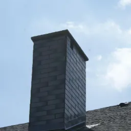 Kominek wykończony ciemnymi płytkami klinkierowymi, widoczny na tle błękitnego nieba i fragmentu dachu pokrytego papą.