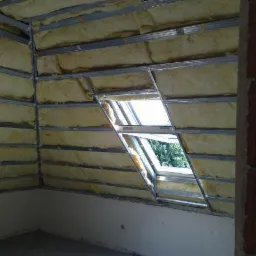 Sufit podwieszany w trakcie montażu z widoczną izolacją z wełny mineralnej i metalową konstrukcją nośną, okno dachowe w skośnej części sufitu przepuszcza światło dzienne.