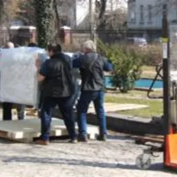 Pracownicy przenoszą zapakowaną maszynę na palecie przy użyciu wózka paletowego na zewnątrz budynku.