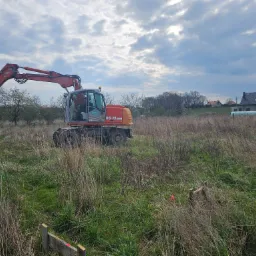 Pomarańczowa koparka kołowa na zarośniętym placu budowy, w tle dom w stanie surowym zamkniętym i pochmurne niebo.