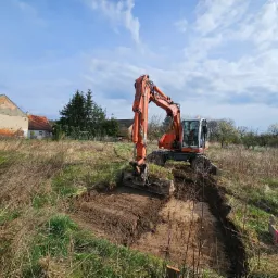 Pomarańczowa koparka wykopuje rów w ziemi na działce z widocznym budynkiem w tle, przy pochmurnym niebie.
