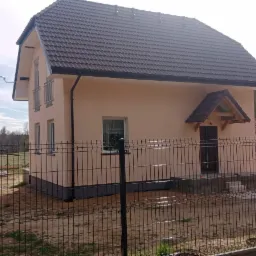 Dwukondygnacyjny dom z beżową elewacją, ciemnym dachem i czarną siatką ogrodzeniową na pierwszym planie, widok z zewnątrz w słoneczny dzień.