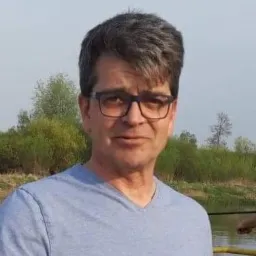 Mężczyzna w okularach i niebieskiej koszulce na tle zieleni i wody. Ujęcie portretowe z naturalnym światłem, widoczne drzewa i fragment rzeki w tle.