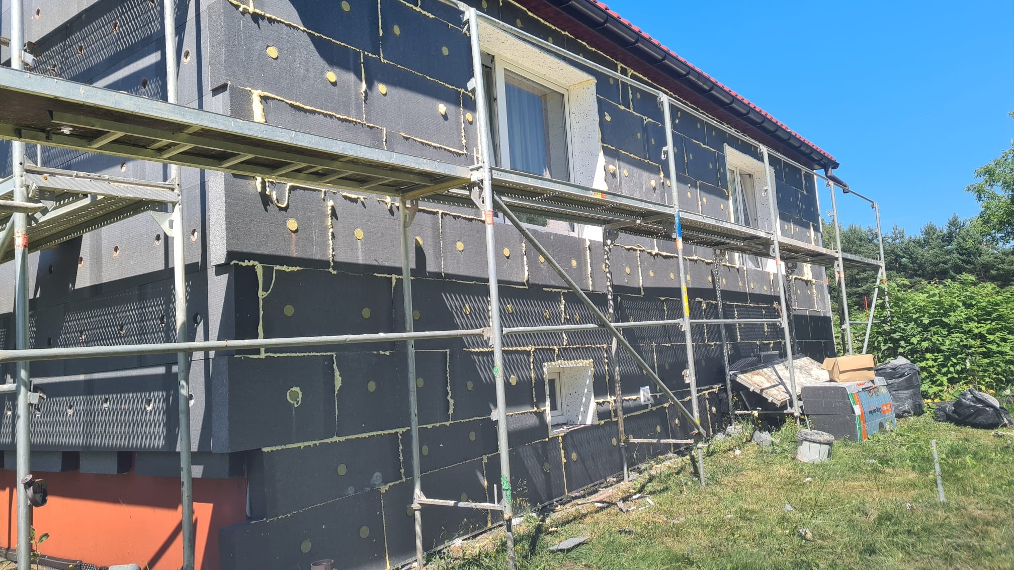 Dom w trakcie ocieplania, widoczne rusztowania i czarne płyty izolacyjne z żółtymi kołkami, częściowo zaklejone pianką montażową. Na trawniku widoczne resztki materiałów budowlanych.