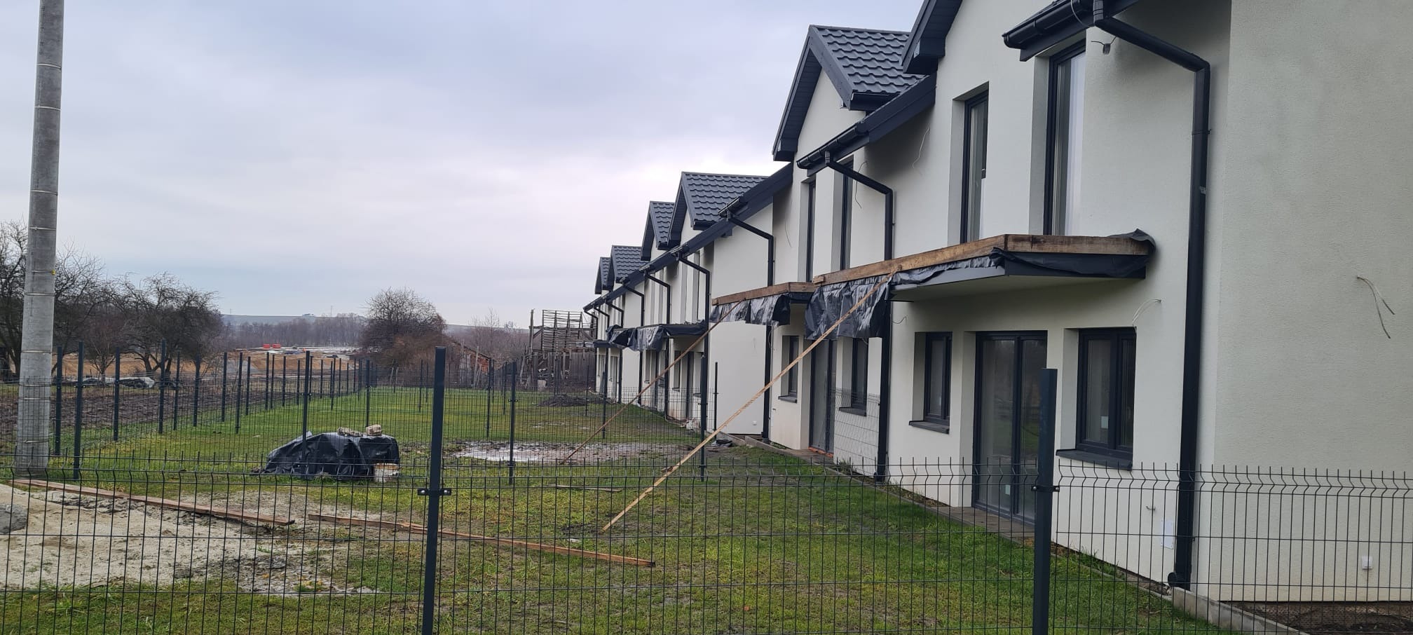 Szereg nowo wybudowanych domów w zabudowie szeregowej z ciemnymi dachami i jasnymi elewacjami, częściowo zabezpieczonych folią, widoczne ogrodzenie z siatki i teren budowy.