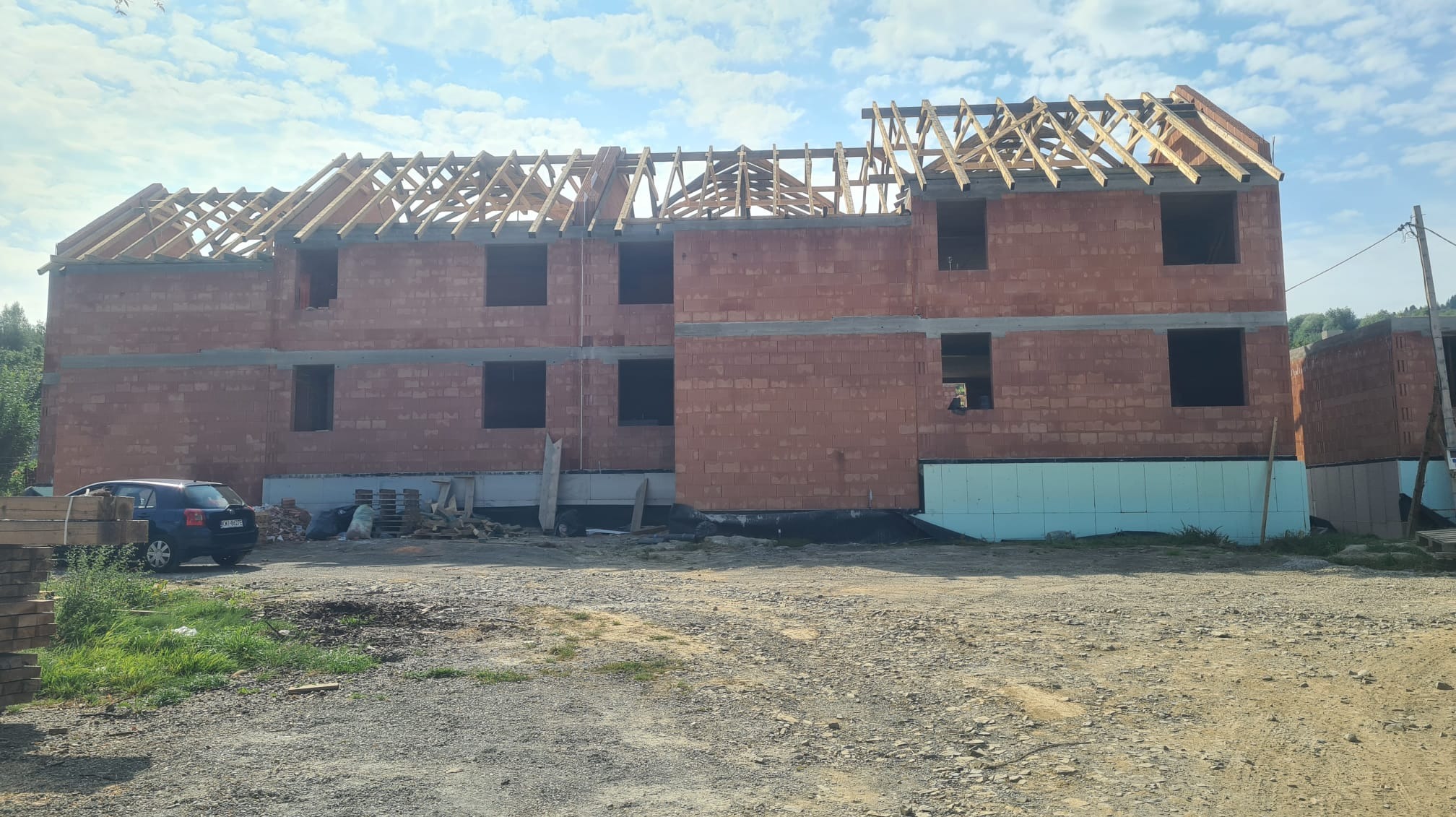 Surowy stan dwukondygnacyjnego domu z czerwonej cegły z widoczną konstrukcją dachu z drewnianych belek, ocieplonym fundamentem i zaparkowanym niebieskim samochodem osobowym z boku.