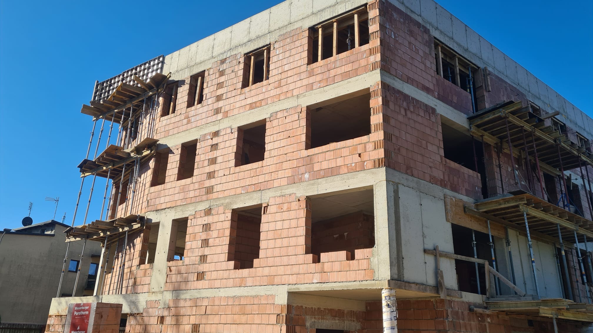 Trzykondygnacyjny budynek w trakcie budowy, z widocznymi pustakami ceramicznymi Porotherm, betonowymi stropami i rusztowaniami na elewacji, w słoneczny dzień.