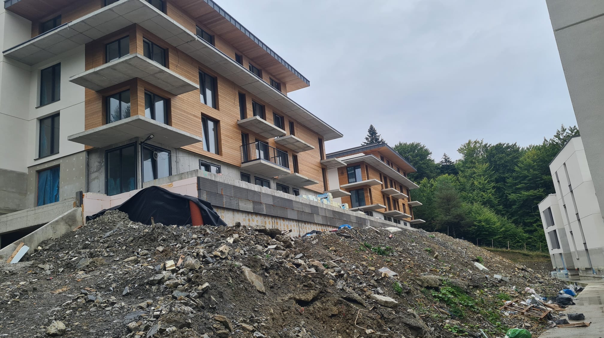 Wielopiętrowy budynek mieszkalny z drewnianą elewacją w trakcie budowy, widok z dołu na plac budowy z hałdą ziemi i gruzu.