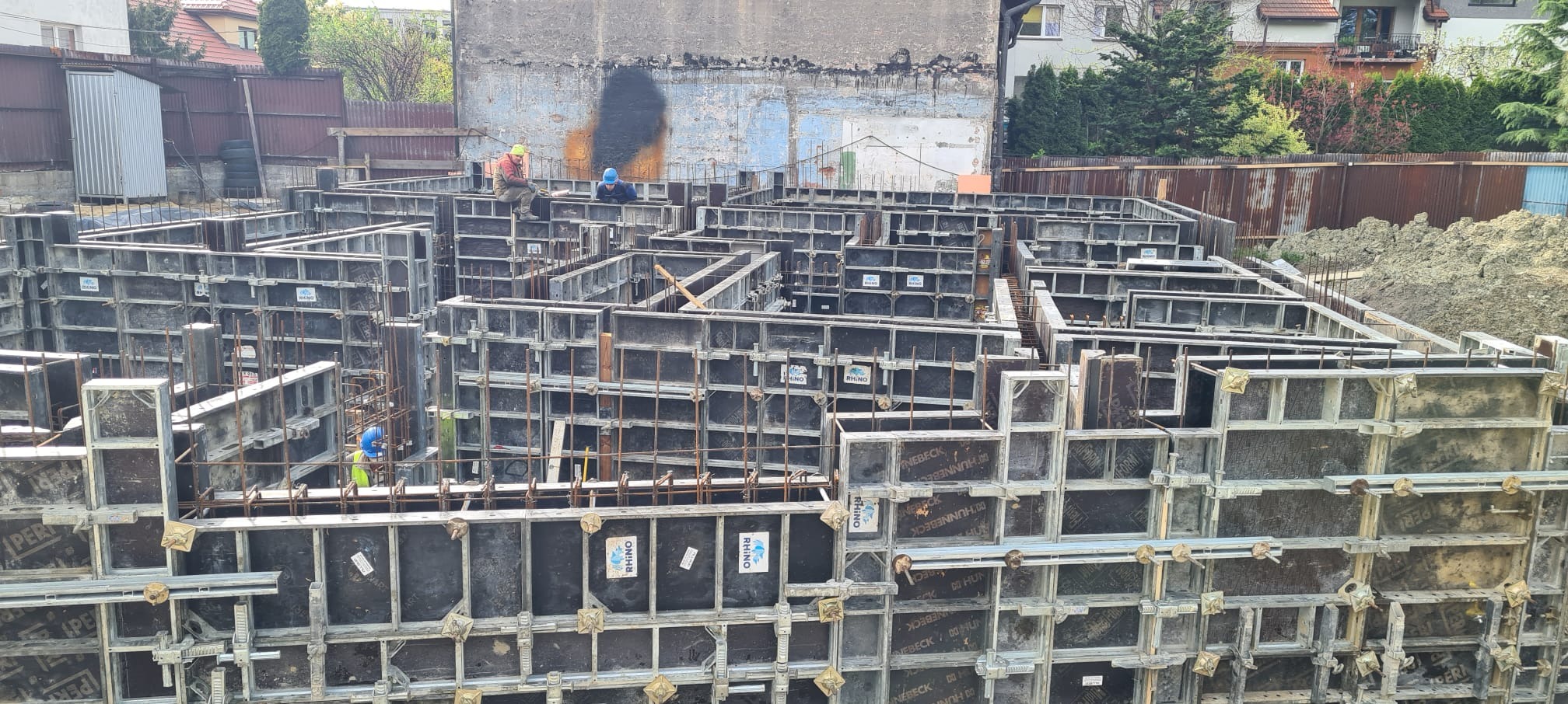 Szalunek fundamentów z widocznymi pracownikami w kaskach, widok z góry na plac budowy z elementami szalunkowymi i zbrojeniami.