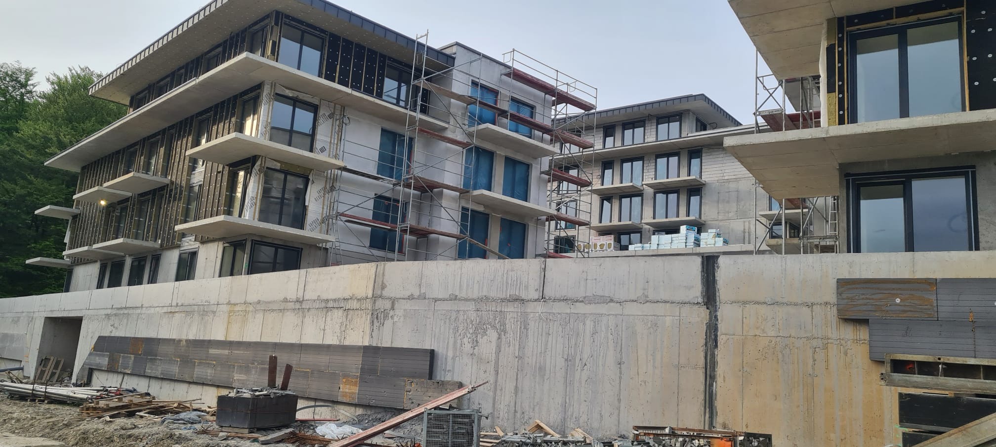 Wielopiętrowy budynek w trakcie budowy z widocznymi rusztowaniami i elementami izolacji termicznej, betonowy fundament i porozrzucane materiały budowlane na pierwszym planie.