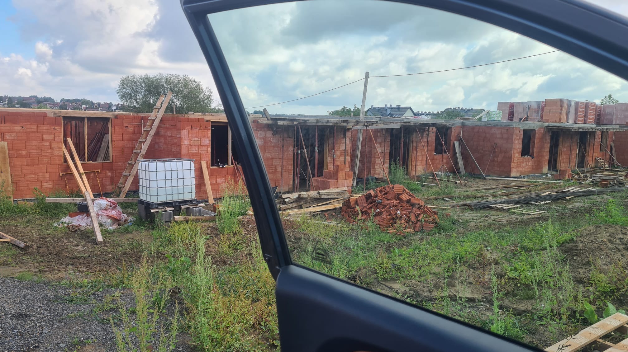 Wznoszone ściany z czerwonej cegły ceramicznej na budowie domu jednorodzinnego, widok przez szybę samochodu, z widocznym pojemnikiem IBC na wodę.