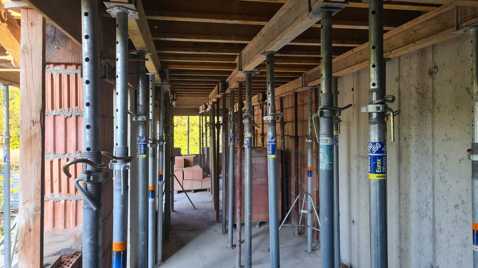 Wnętrze budowy domu z widocznymi ścianami z pustaków ceramicznych, drewnianym szalunkiem stropu podpartym metalowymi stemplami z widocznymi etykietami Eurex i Doka, na podłodze ułożone pustaki.