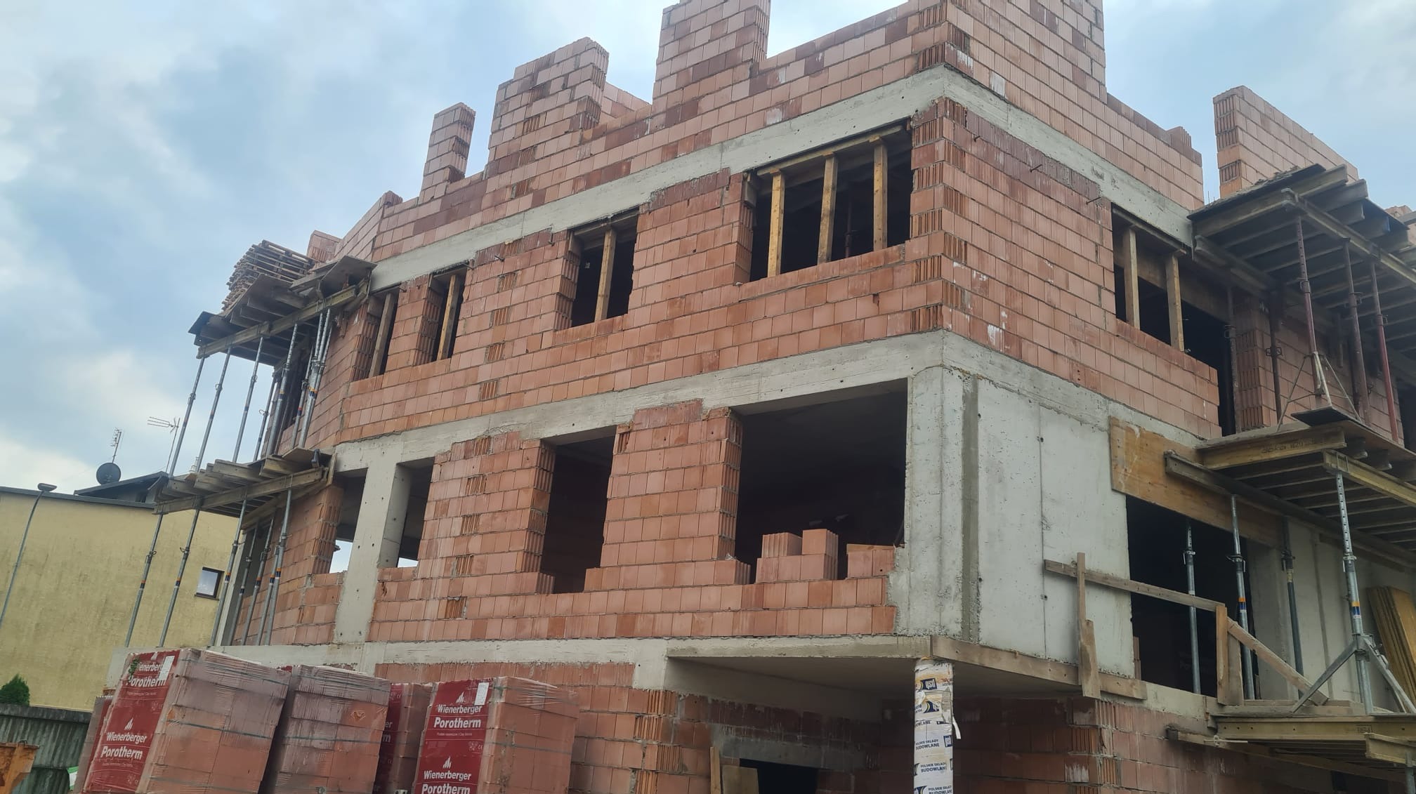 Wznoszenie dwukondygnacyjnego budynku z czerwonej cegły Porotherm, widoczne betonowe elementy konstrukcyjne i rusztowania, palety z cegłą u podstawy.