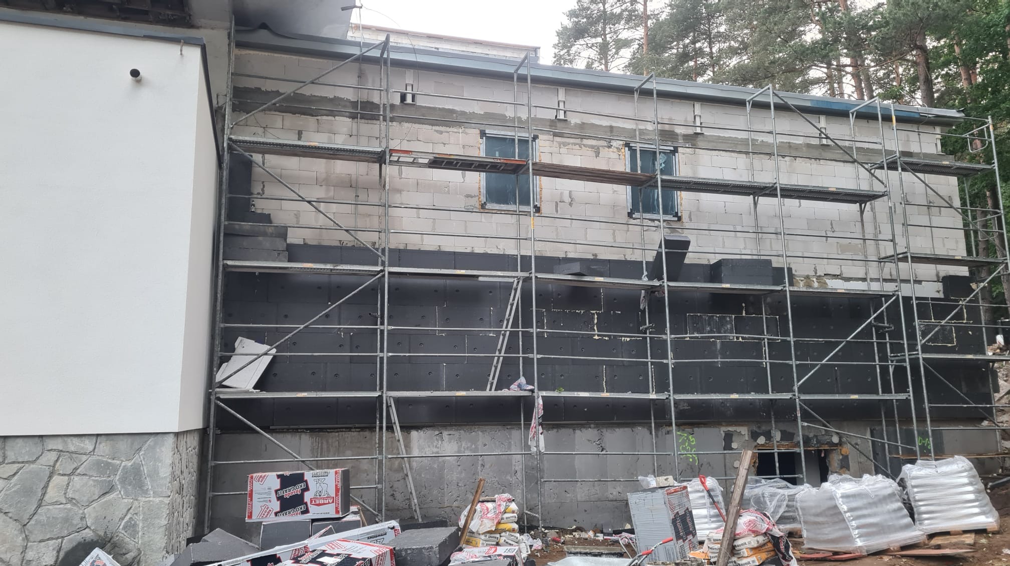 Budynek w trakcie ocieplania: ściana z bloczków, częściowo pokryta czarnym styropianem, rusztowanie na całej wysokości, materiały budowlane u podstawy ściany.