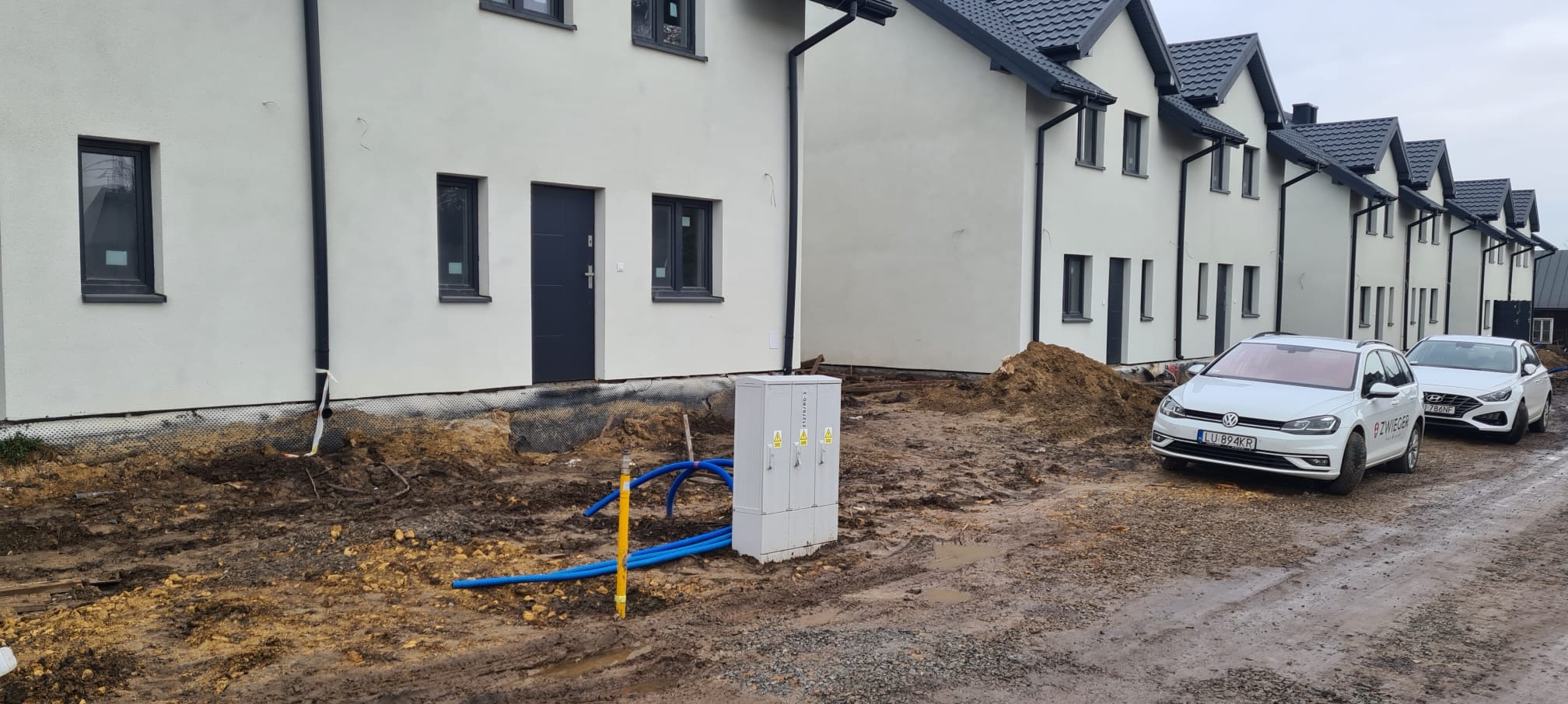 Szereg białych domów w trakcie budowy, widoczne przyłącza mediów i dwa samochody na błotnistej drodze.