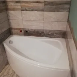 Narożna wanna obudowana białą zabudową, otoczona płytkami imitującymi drewno na ścianach i podłodze. Widoczne niepodłączone przyłącza wodne.