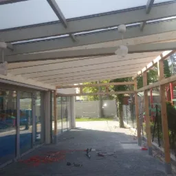 Konstrukcja drewnianej pergoli w trakcie budowy, widoczne narzędzia i kable na brukowanej posadzce, przeszklone drzwi przesuwne w tle.