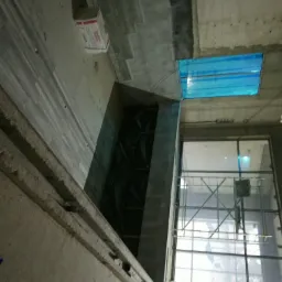 Surowy stan wnętrza domu w trakcie budowy: widoczne szare ściany, okno zabezpieczone folią, rusztowania na zewnątrz i karton na podłodze.