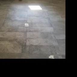 Ułożona podłoga z szarych, matowych płytek w łazience, z odpływem liniowym z motywem kwiatowym, widoczne rury instalacji sanitarnej przy ścianie.