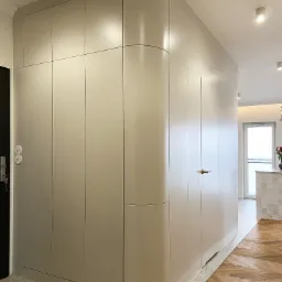 Beżowa szafa na wymiar z zaokrąglonym narożnikiem, wkomponowana w korytarz z parkietem w jodełkę i terazzo na podłodze.