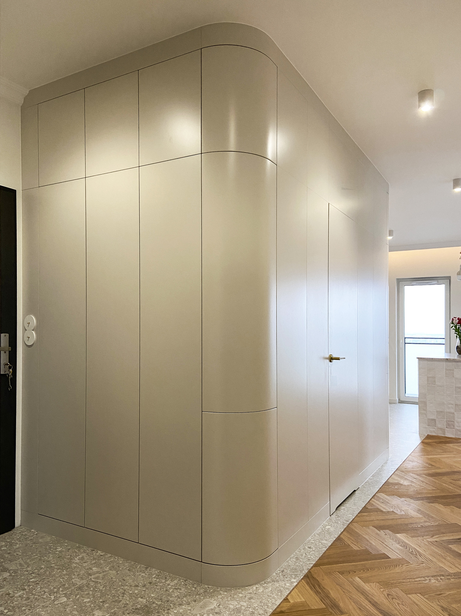 Beżowa szafa na wymiar z zaokrąglonym narożnikiem, wkomponowana w korytarz z parkietem w jodełkę i terazzo na podłodze.