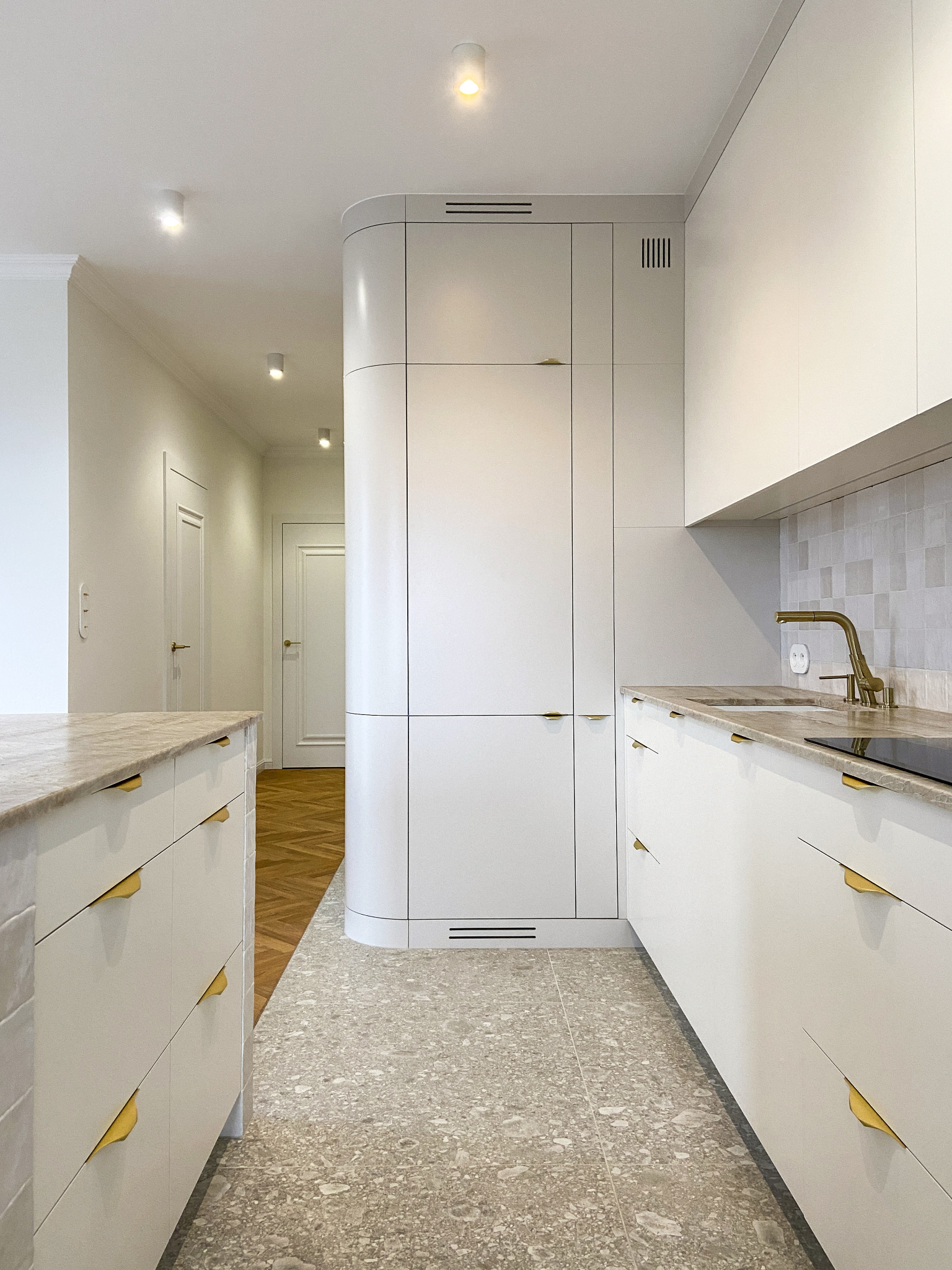 Jasna kuchnia z białymi frontami i złotymi uchwytami, marmurowy blat, zaokrąglona zabudowa spiżarni, podłoga w terazzo, widok na przedpokój z parkietem.