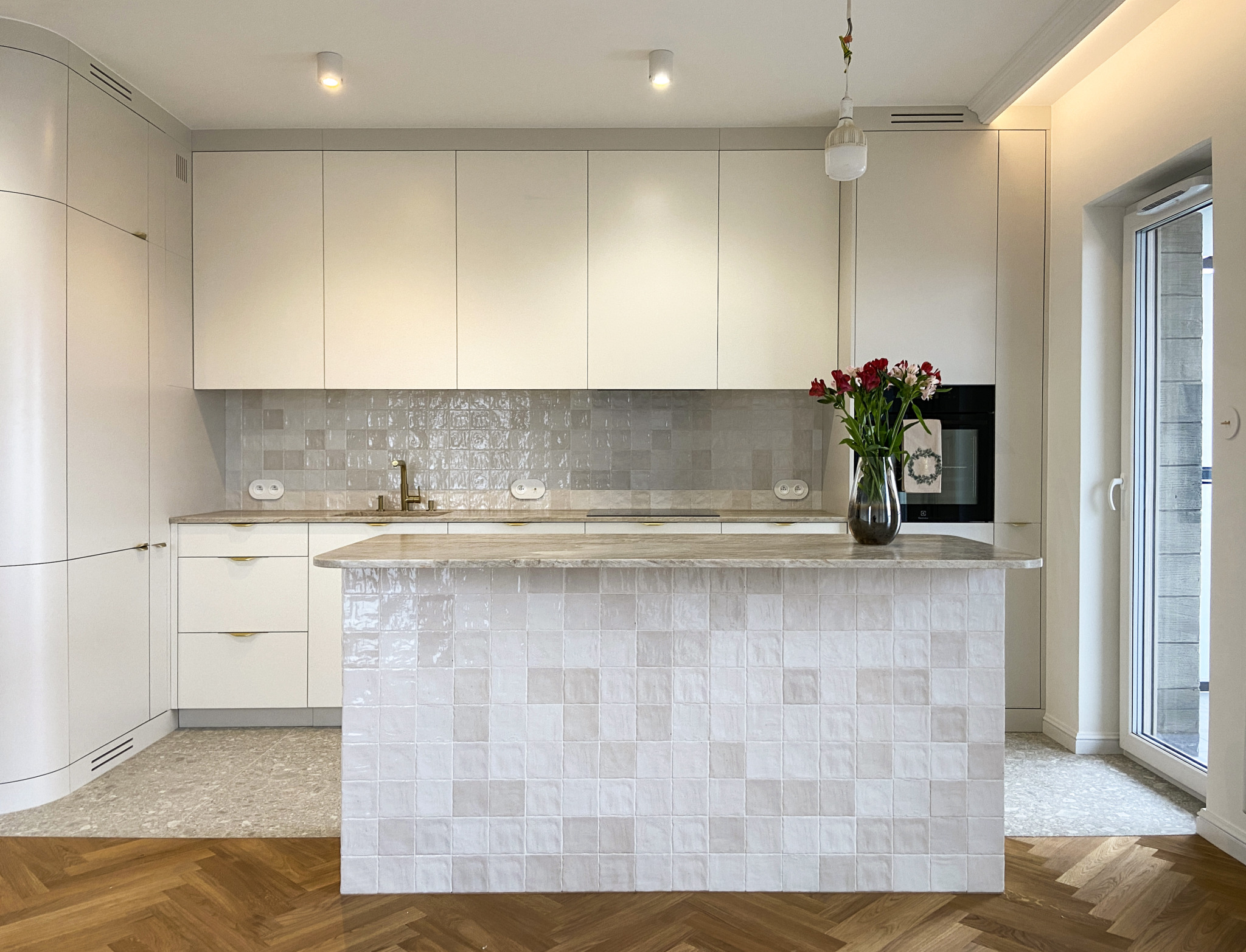 Jasna kuchnia z wyspą wykończoną błyszczącymi, kwadratowymi płytkami, parkiet w jodełkę i bukiet czerwonych kwiatów w szklanym wazonie na blacie.