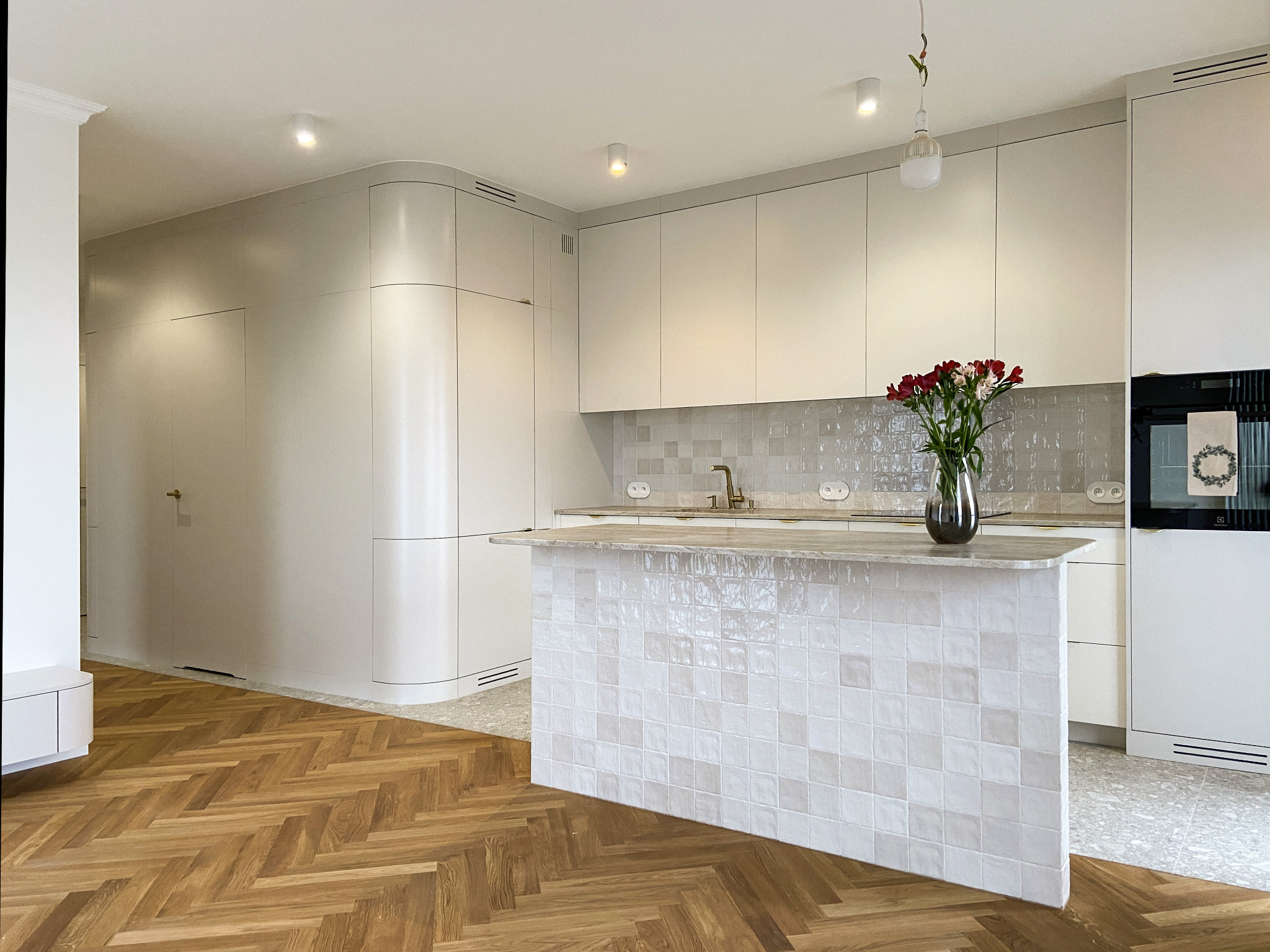 Jasna, nowoczesna kuchnia z wyspą wykończoną kwadratowymi płytkami, parkiet w jodełkę, czerwone kwiaty w wazonie na blacie.