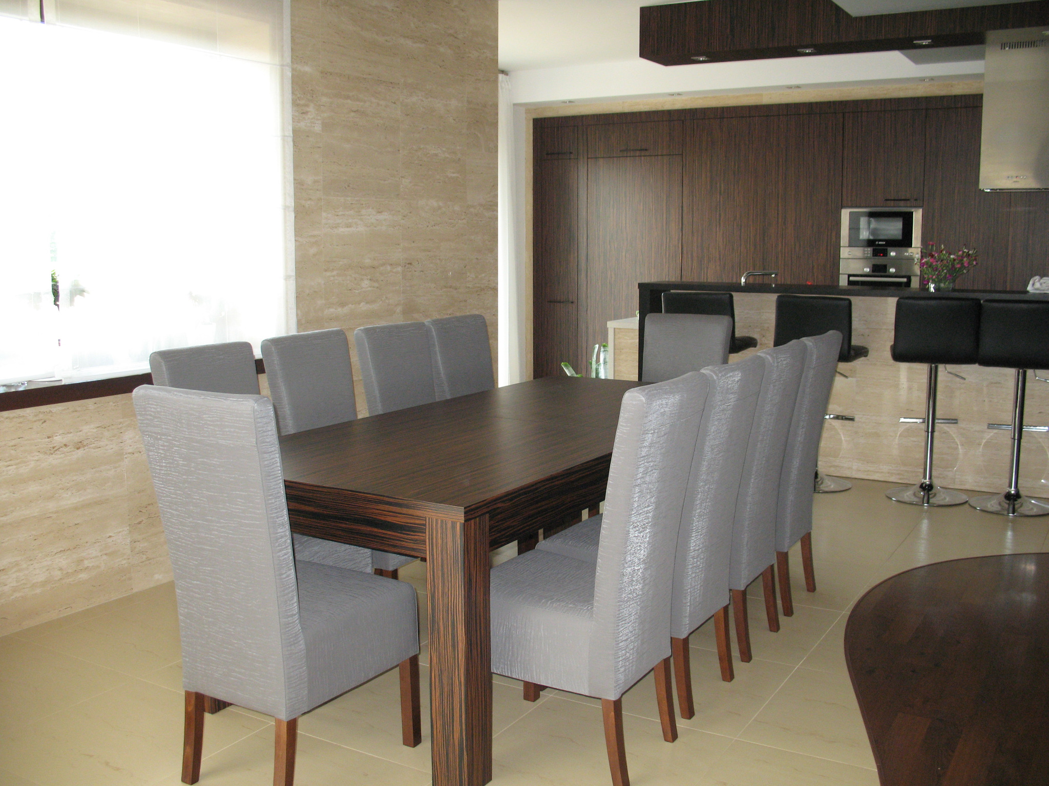 Nowoczesna jadalnia z ciemnym stołem z wyraźnym usłojeniem i ośmioma szarymi krzesłami z drewnianymi nogami na tle kuchni z ciemnymi szafkami i barkiem.