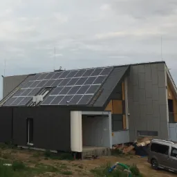 Nowoczesny dom z panelami słonecznymi na dachu, częściowo wykończony szarymi panelami elewacyjnymi, z widoczną konstrukcją w stanie surowym w niektórych miejscach, zaparkowany samochód dostawczy...