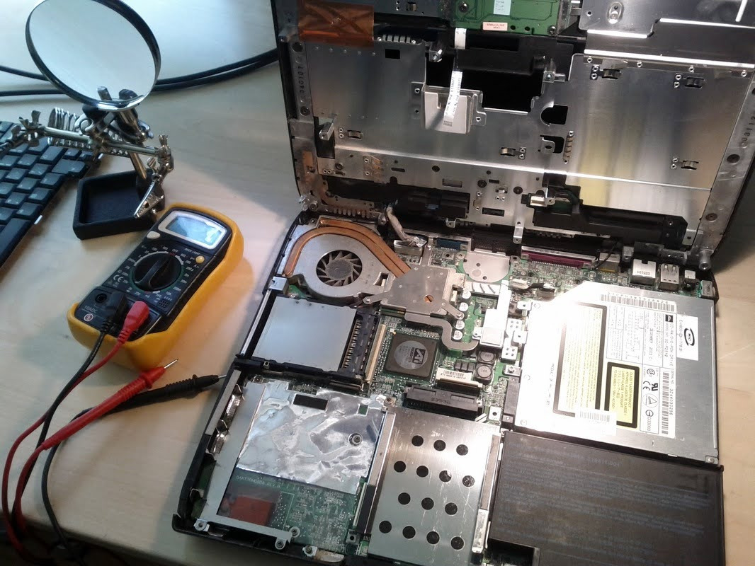 Otwarty laptop z widocznymi podzespołami wewnętrznymi, obok multimetr z podłączonymi przewodami pomiarowymi, lupa na statywie i fragment klawiatury na biurku.