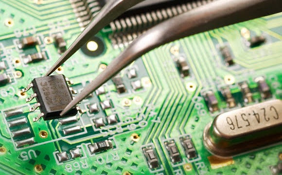 Precyzyjne umieszczanie układu scalonego na zielonej płycie głównej za pomocą metalowej pęsety, widoczne liczne komponenty elektroniczne.