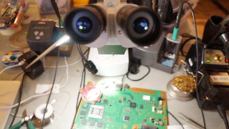 Płyta główna konsoli do gier pod mikroskopem, otoczona narzędziami serwisowymi i elementami elektronicznymi na warsztatowym stole.