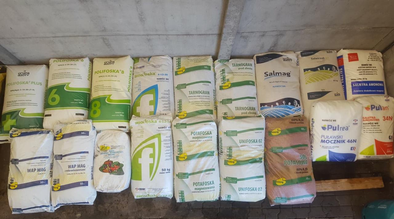 Różne worki z nawozami, ułożone w rzędach pod betonową ścianą, widoczne nazwy producentów i typy nawozów: Polifoska, Tarnogran, Salmag, Pulrea. Część worków jest biała, część ma zielone i brązowe...
