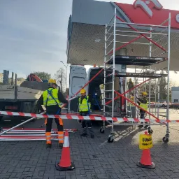 Pracownicy w odblaskowych kamizelkach i kaskach montują rusztowanie przy stacji benzynowej, przygotowując się do prac na dachu, widoczny samochód dostawczy i tablica ostrzegawcza 'Remont dachu'.
