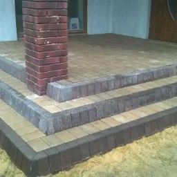 Schody wejściowe z kostki brukowej dwukolorowej, wykończone ciemną obwódką, prowadzące do drzwi wejściowych. Filar z cegły po lewej stronie.