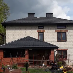 Montaż ciemnoszarej dachówki na dwupiętrowym domu z cegły, widoczne rusztowania i pracownik w trakcie układania, z pochmurnym niebem w tle.