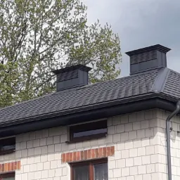 Szary dach z dwoma kominami wentylacyjnymi, widok z dołu na tle drzewa i pochmurnego nieba, biała ceglana ściana budynku z ciemnymi oknami.