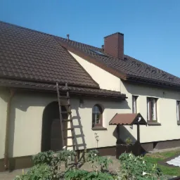 Dom jednorodzinny z brązową dachówką, drabina oparta o ścianę, widoczne elementy ocieplenia na dachu i ścianach, studzienka zadaszona na pierwszym planie.