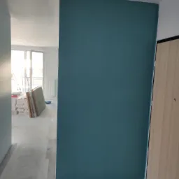 Świeżo pomalowana ściana na ciemny turkusowy kolor w nowym mieszkaniu, widoczne kartony z panelami i drabina w tle, surowy beton na podłodze.