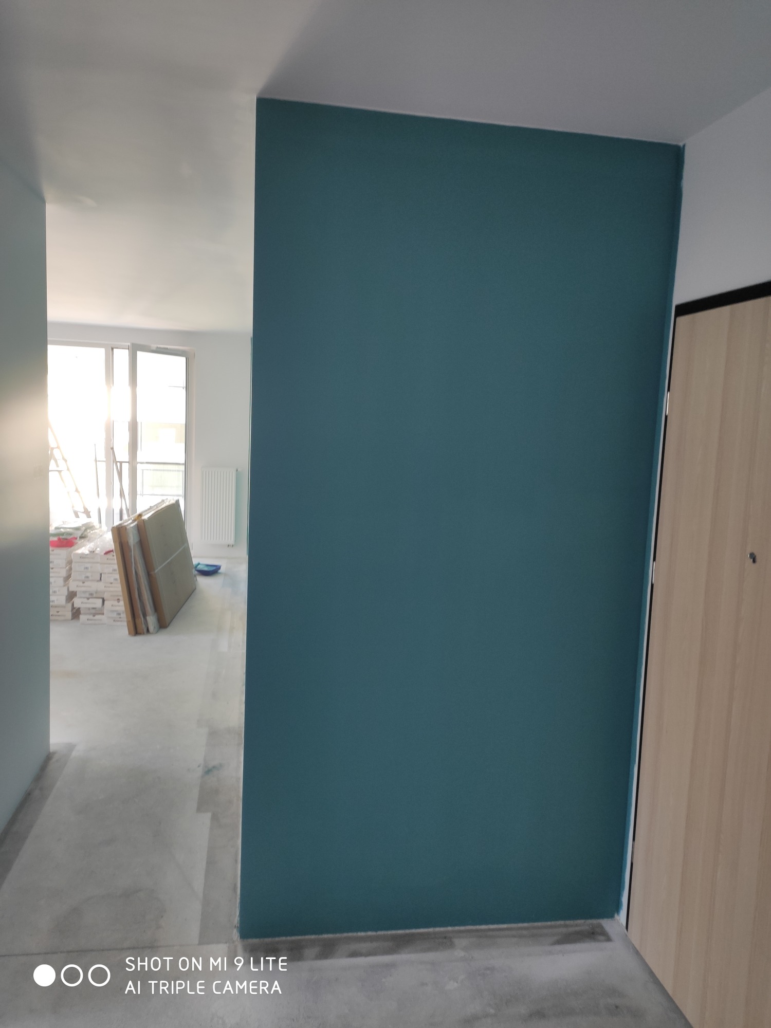 Świeżo pomalowana ściana na ciemny turkusowy kolor w nowym mieszkaniu, widoczne kartony z panelami i drabina w tle, surowy beton na podłodze.