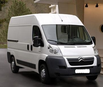 Biały samochód dostawczy typu furgon marki Citroen, zaparkowany przed budynkiem z widocznym zadaszeniem i oświetleniem zewnętrznym.