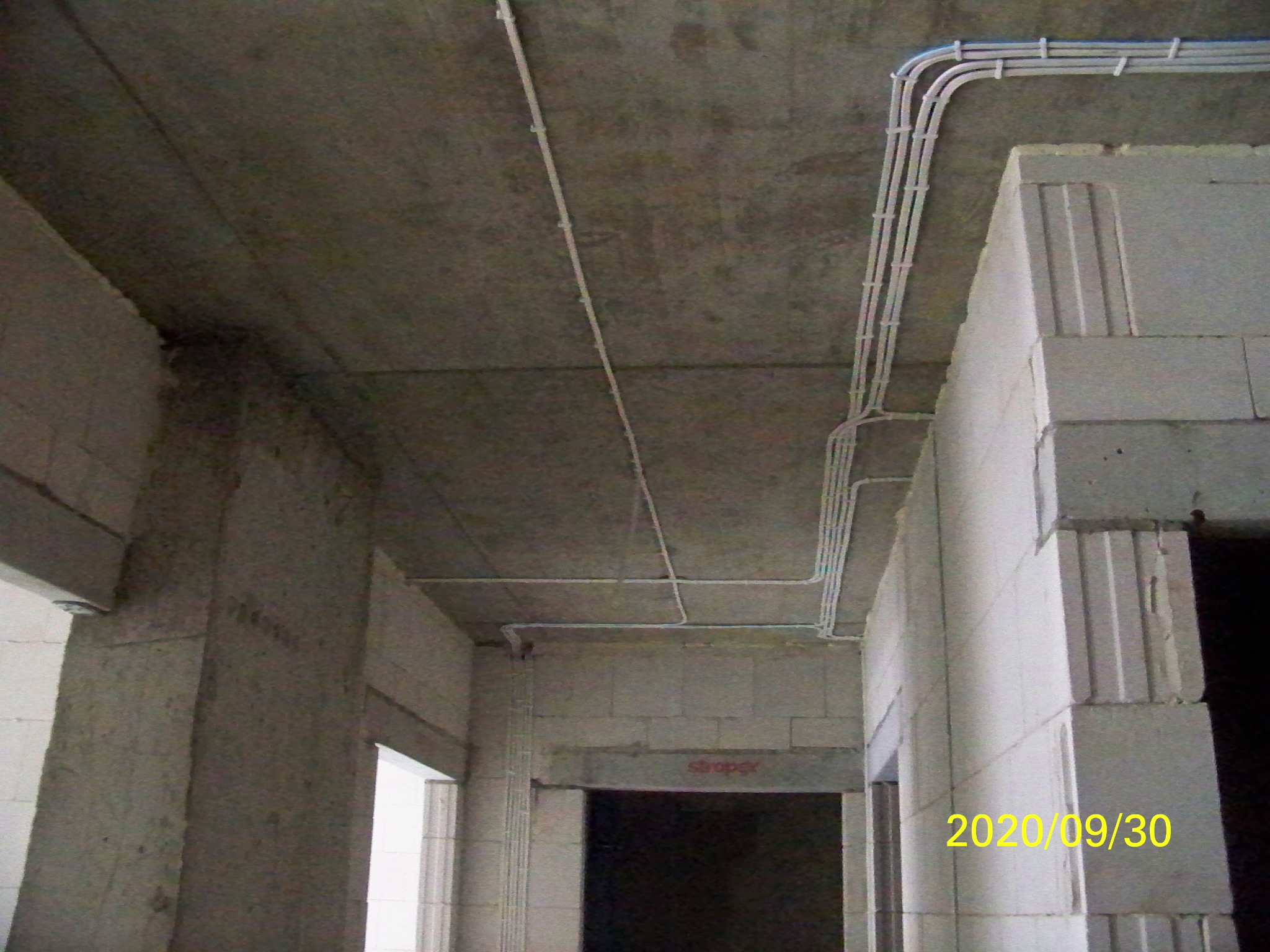 Surowe wnętrze w trakcie budowy z widocznymi, prowadzonymi po suficie i ścianach rurami instalacji elektrycznej, zamocowanymi za pomocą uchwytów. Data 2020/09/30 widoczna w prawym dolnym rogu.