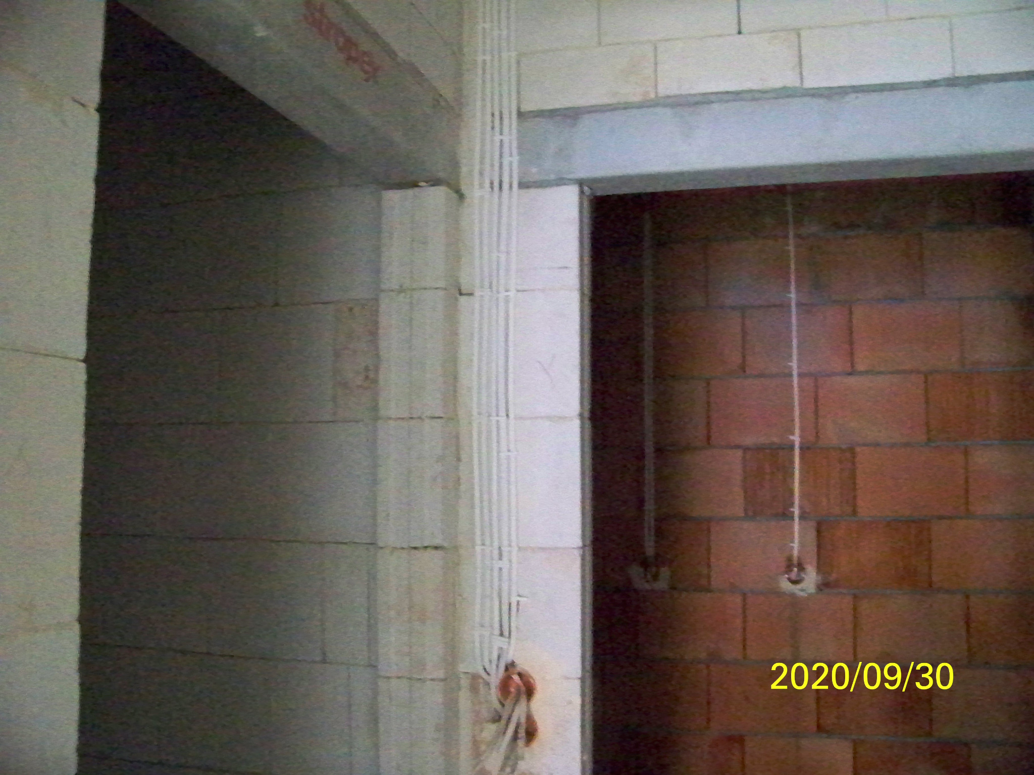 Wiązka białych kabli elektrycznych prowadzona w peszlach, zamocowana na ścianie z białych bloczków, w tle widoczna ściana z czerwonej cegły, zdjęcie z datą wykonania w prawym dolnym rogu.