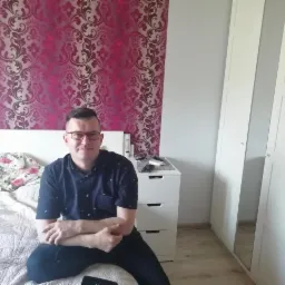 Mężczyzna w okularach siedzi na łóżku z wzorzystą pościelą na tle ściany w bordowe ornamenty. Obok biała szafa i komoda z szufladami.