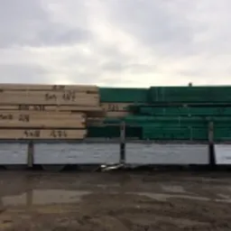 Transport tarcicy na platformie ciężarówki, część pakietów owinięta zieloną folią, widoczne oznaczenia wymiarów i ilości na drewnie.