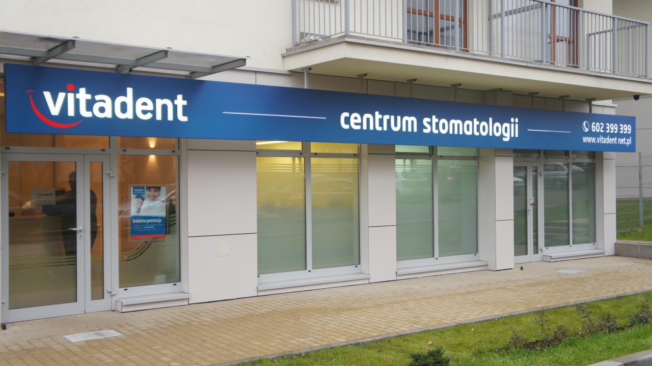 Zewnętrzny widok na budynek centrum stomatologii Vitadent z dużym, podświetlanym szyldem w kolorze niebieskim z białym napisem i czerwonym elementem graficznym.