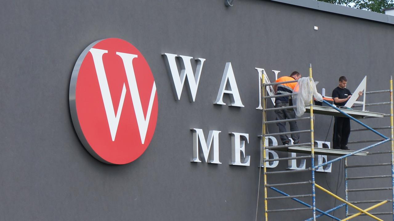 Montaż przestrzennego logo 'WANN MEBLE' na szarej fasadzie budynku. Dwóch monterów na rusztowaniu przykłada białe litery do ściany.