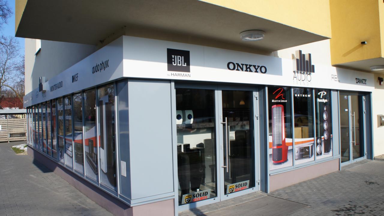 Fasada sklepu audio z logo JBL, Onkyo, Audio Trendt i innymi markami. Widoczne plakaty z głośnikami w oknach. Szary budynek z białymi napisami.