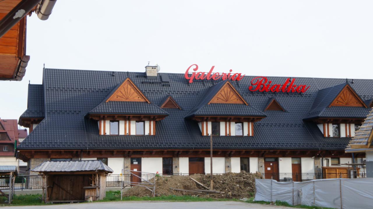 Budynek w stylu regionalnym z ciemnym dachem i czerwonym napisem 'Galeria Białka' na górze. Widoczne drewniane elementy dekoracyjne i mała budka z boku.